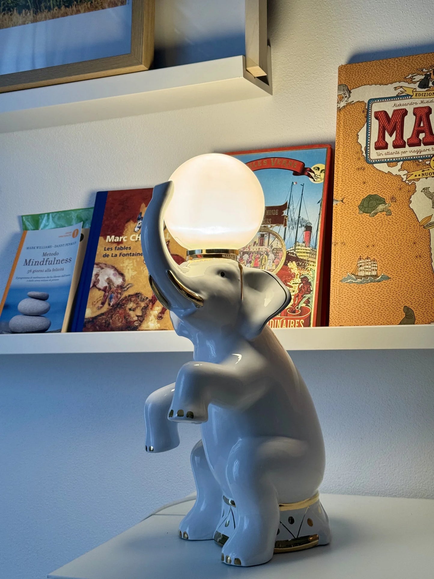 Lampada in ceramica bianca a forma di elefante – accesa vicina a una mensola con libri per bambini