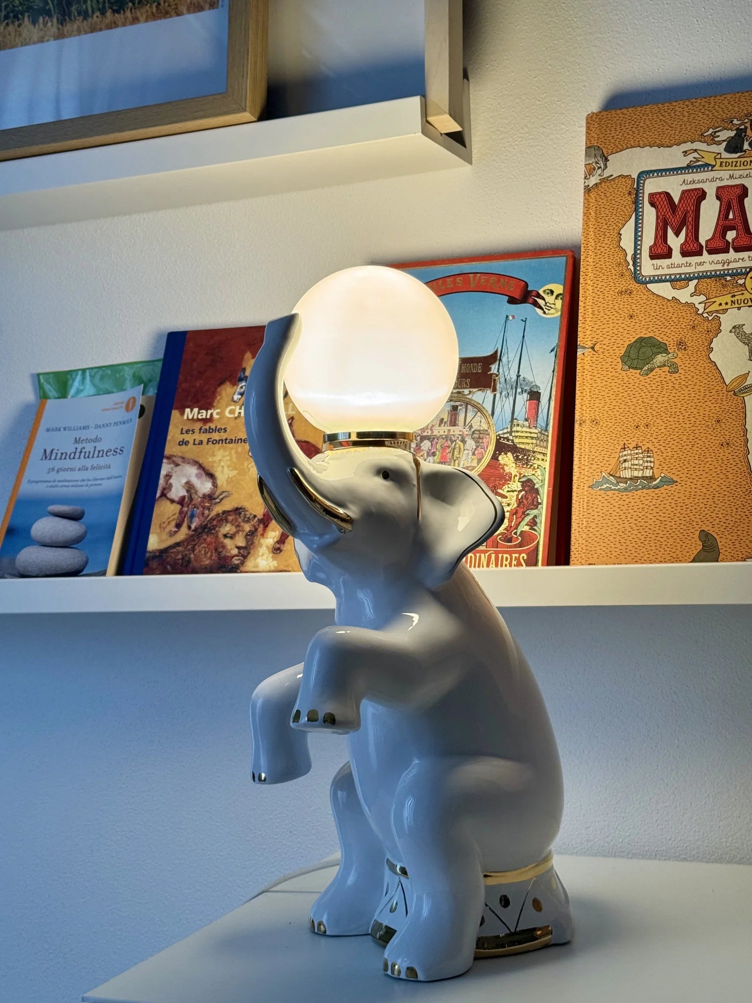 Lampada in ceramica bianca a forma di elefante – accesa vicina a una mensola con libri per bambini