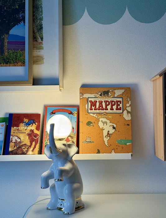 Lampada in ceramica bianca a forma di elefante – accesa sopra uno scaffale bianco con libri